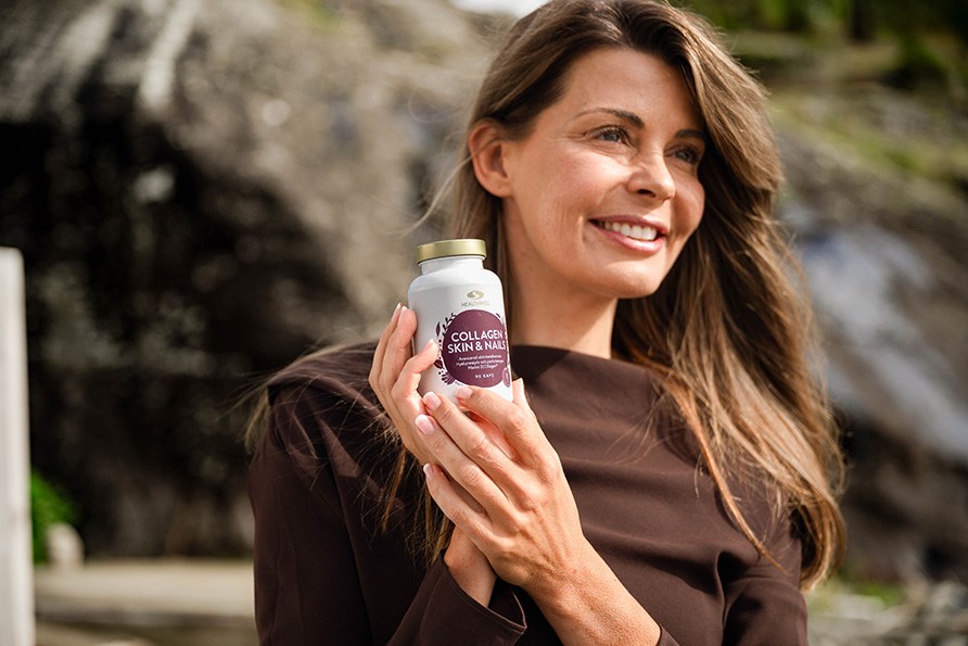 Grinende kvinde med langt brunt hår og brun trøje holder op en bøtte Healthwell Collagen Skin & Nails udendørs, foran en klippe.