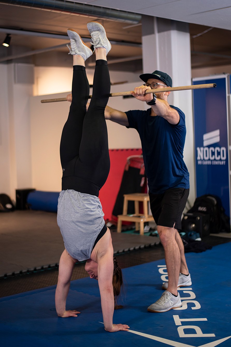 En kvinde står på hænder op af en pind, som holdes af en mand, i et træningslokale. En kvinde står på hænder op af en pind, som holdes af en mand, i et træningslokale.