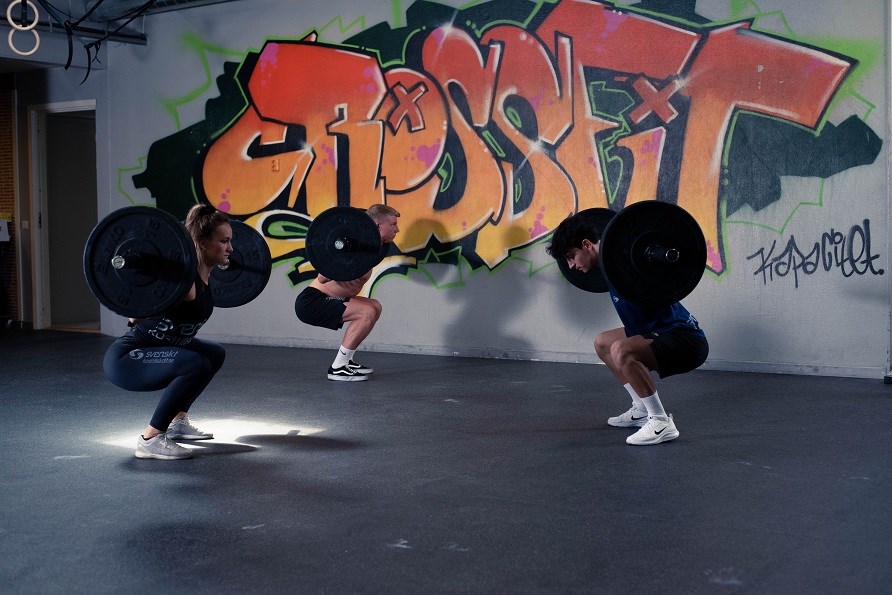 En kvinde og to m�nd i et fitnesscenter laver squat med en stang p� ryggen,