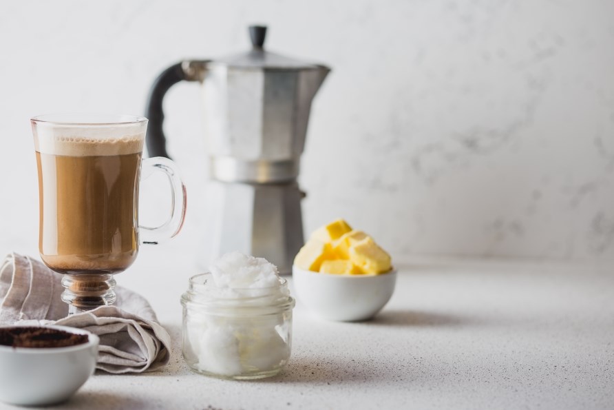En kop kaffe str ved siden af en kop med kokosolie, en kop smr og en fransk kaffepres.