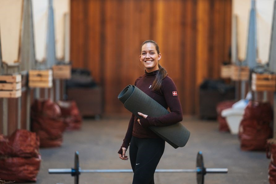 Cathrine Laudrup Dufour står med en træningsmåtte og smiler foran en vægtstang. Cathrine Laudrup Dufour står med en træningsmåtte og smiler foran en vægtstang.