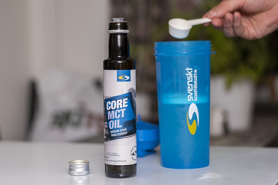Billede af Core MCT-olie i flaske, en shaker i baggrunden og en hnd, der holder en 1 spsk MCT-olie i shakeren.