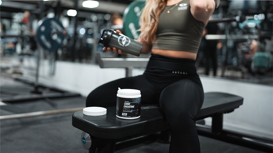En kvinde i et tr�ningsmilj� sidder p� en tr�ningsb�nk med en b�tte Core Creatine Pro foran sig.