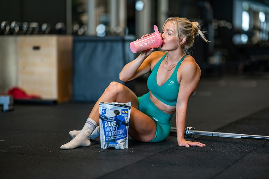 Kvinde sidder p gulvet i et fitnesscenter og drikker Core Whey Protein fra en shaker.