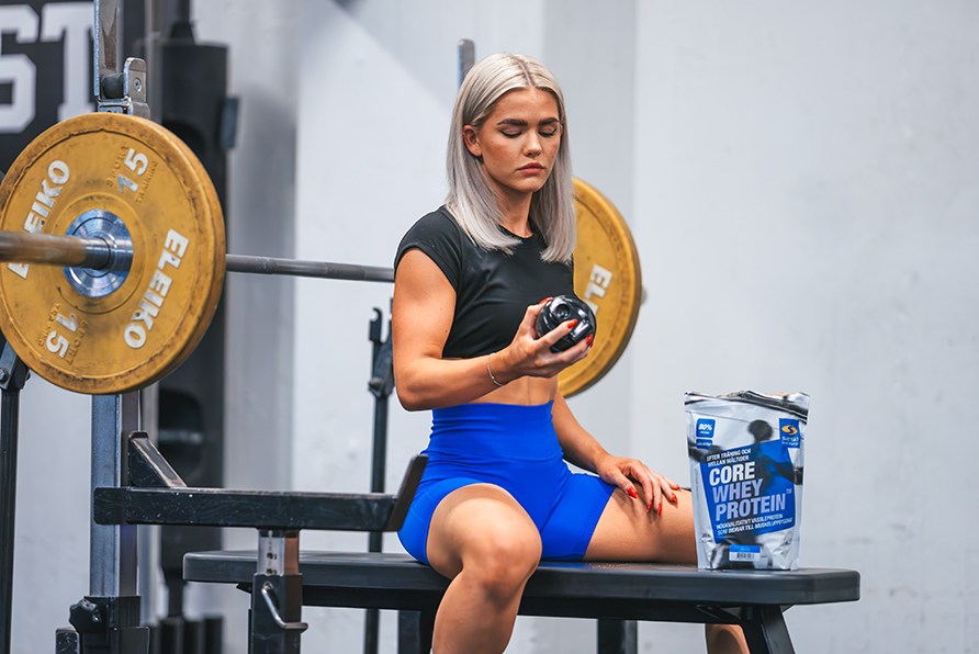 Kvinde sidder på bænk og holder shaker i et fitnesscenter. Kvinde sidder på bænk og holder shaker i et fitnesscenter.