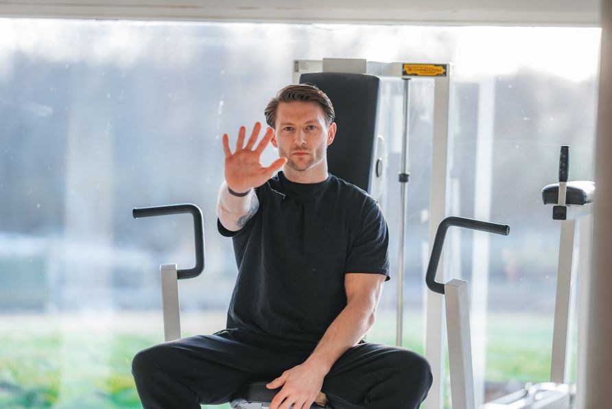Kenneth rækker hånden oppe med fem fingre, siddende på en maskine i et træningscenter. Kenneth rækker hånden oppe med fem fingre, siddende på en maskine i et træningscenter.